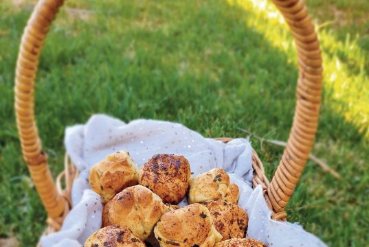 A basket of golden baked small round rolls with flecks of dried herbs on them, The basket is sat on green grass.