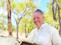 Smiling man in protective bee keeping attire holding a tray from a bee hive. The tray is covered in honeycomb and bees.