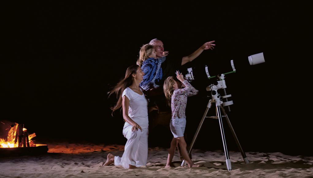 A mum, dad and two daughters on a beach with a telescope pointed skyward. The dad is pointing to the night sky and a campfire burns behind them.