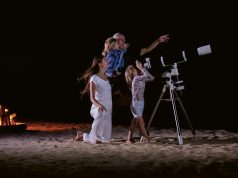 A mum, dad and two daughters on a beach with a telescope pointed skyward. The dad is pointing to the night sky and a campfire burns behind them.