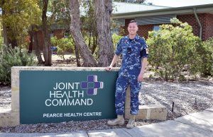 A young air force officer in blue camouflage uniform in front of a sign that says Joint Health Command Pearce Health Centre.
