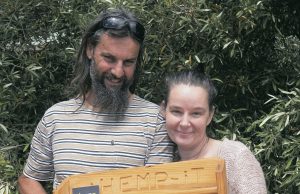A man with dark long hair and beard and smiling woman holding a wooden display rack with tins of hemp products Pain Balm and Save Your Skin