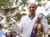 A beaming man in a white jacket holds a newborn dwarf goat in his left hand and a large, silver trophy cup in his right