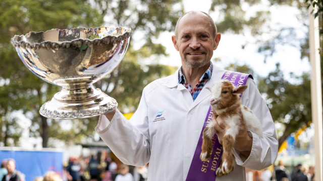A beaming man in a white jacket holds a newborn dwarf goat in his left hand and a large, silver trophy cup in his right