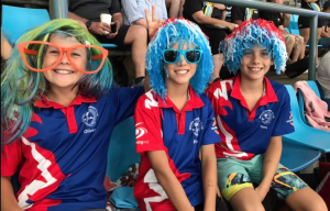 Three smiling boys in blue and red Moora Swim Club uniforms, oversized novelty glasses and wigs